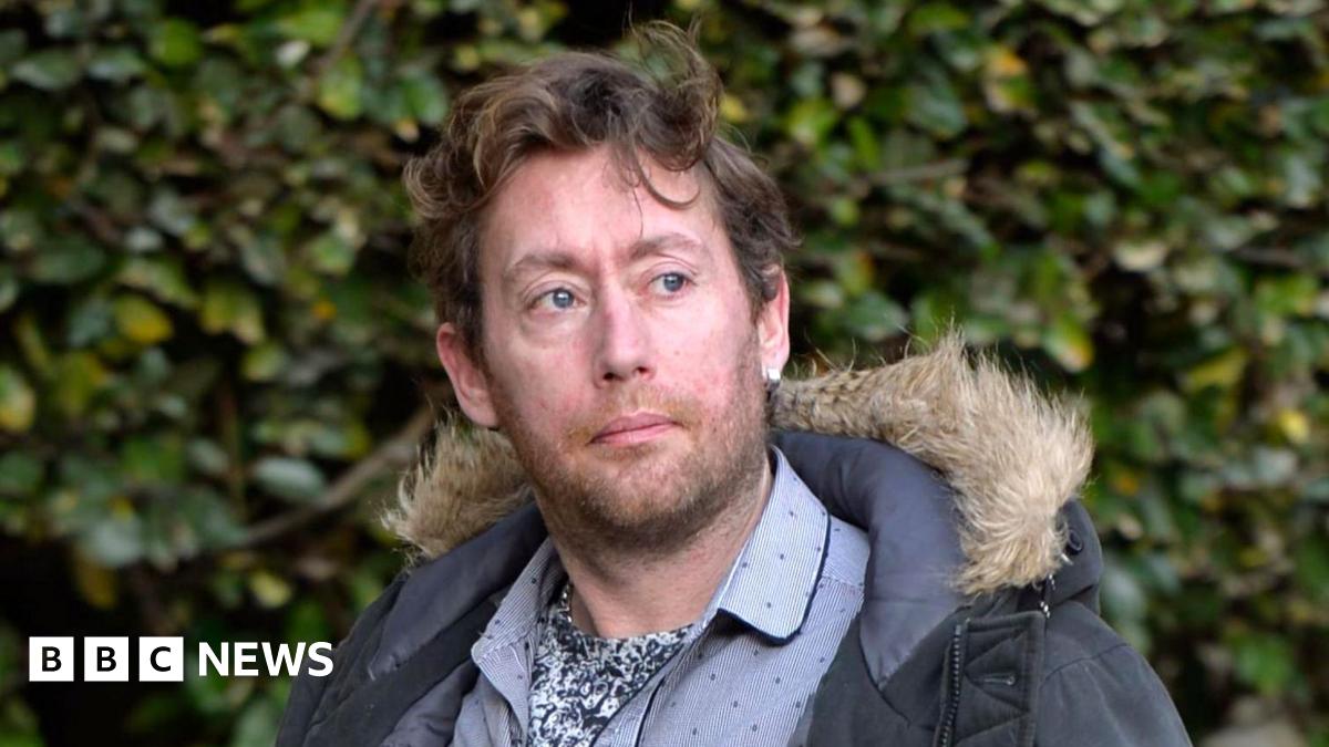 A man in his late 30s with wavy brown hair, a blue parka and a blue shirt outside the High Court in Glasgow. There are bushes in the background of the image and he looks to the right of where the camera is.