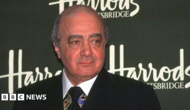 Mohamed Al Fayed in a suit stood in front of a sign that says Harrods.