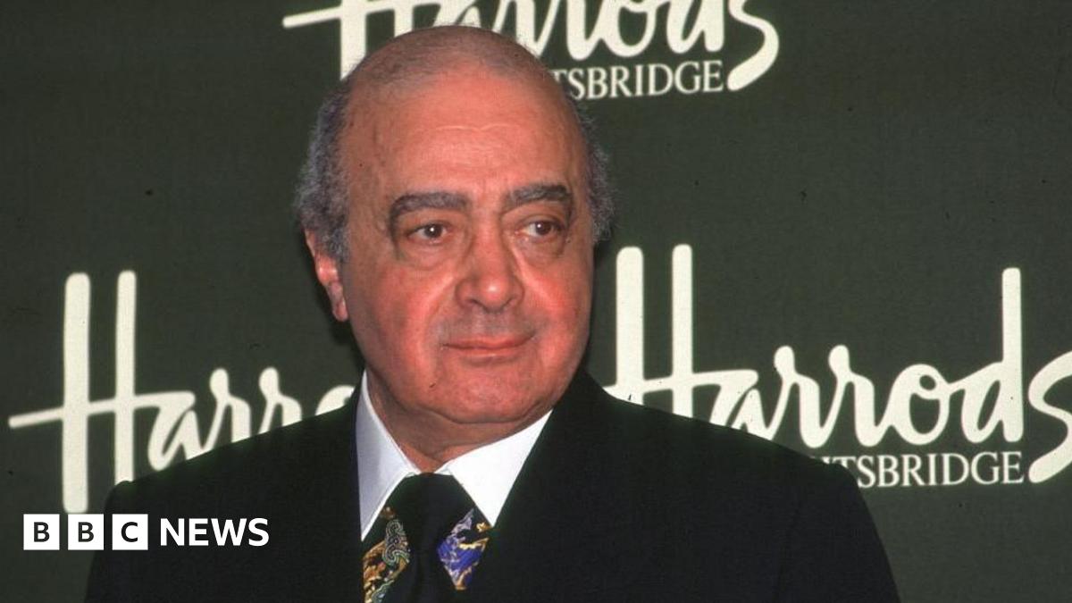 Mohamed Al Fayed in a suit stood in front of a sign that says Harrods.