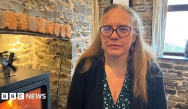 Sproson-Jones is photographed looking into the camera with a serious expression. She is wearing a black blouse with white and green flowers, and a black cardigan. Her hair is long and blonde, and is tied back in a half-up hairstyle. She's also wearing a pair of dark-framed glasses. She's sat next to a black log burner, which is lit and has flames inside. The walls of the pub and fireplace are exposed grey stone, with a window behind her.