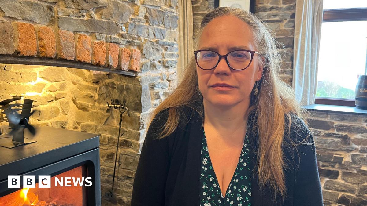 Sproson-Jones is photographed looking into the camera with a serious expression. She is wearing a black blouse with white and green flowers, and a black cardigan. Her hair is long and blonde, and is tied back in a half-up hairstyle. She's also wearing a pair of dark-framed glasses. She's sat next to a black log burner, which is lit and has flames inside. The walls of the pub and fireplace are exposed grey stone, with a window behind her.