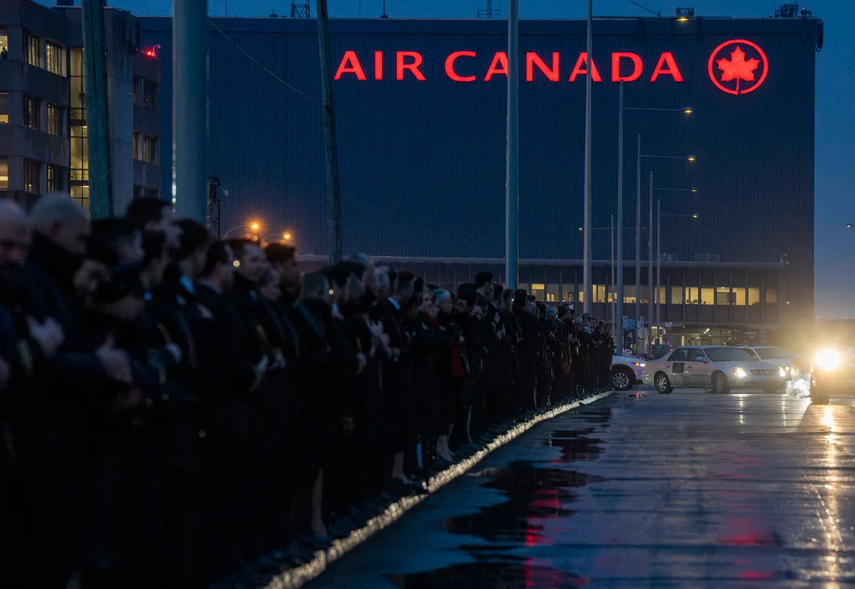 Pilots in uniform line a street on a rainy day at dusk as a funeral car home turns the corner in the distance