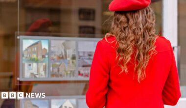 Woman wearing bright red beret and red coat faces an estate agent's window advertising homes for sale