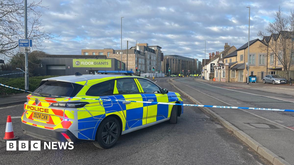 The rear of a police car is shown inside police tape on a road. In the distance, more police cars and officers can be seen