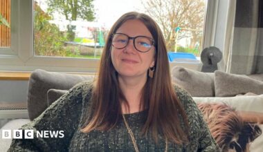 Amie Watson, sat on her sofa, looking into the camera. She is wearing an olive green cable knit jumper, and a long gold necklace. She has long brown hair tucked behind her ears, and a pair of tortoiseshell-framed glasses. She's sat on a grey sofa with a cushion with a picture of a Highland cow on it. Behind her is a large window, and you can see into the garden where there's a trampoline and a climbing frame.