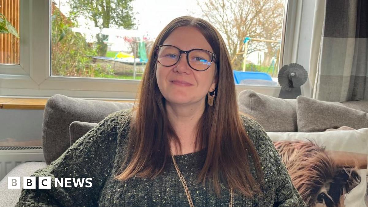 Amie Watson, sat on her sofa, looking into the camera. She is wearing an olive green cable knit jumper, and a long gold necklace. She has long brown hair tucked behind her ears, and a pair of tortoiseshell-framed glasses. She's sat on a grey sofa with a cushion with a picture of a Highland cow on it. Behind her is a large window, and you can see into the garden where there's a trampoline and a climbing frame.