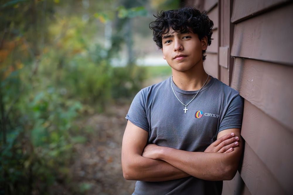 Teen boy with curly hair wearing a gray t-shirt and cross necklace stands with arms crossed by a wooden wall, with greenery in the background.