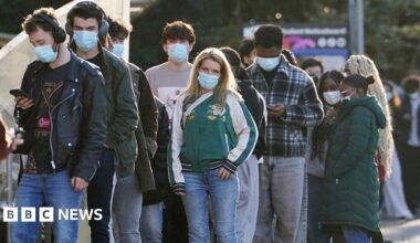 Students wait in line at the entrance to the sports hall at University of Kent campus in Canterbury. They are wearing masks and coats. Some are looking their phones, one has headphones on. Some are chatting in groups.