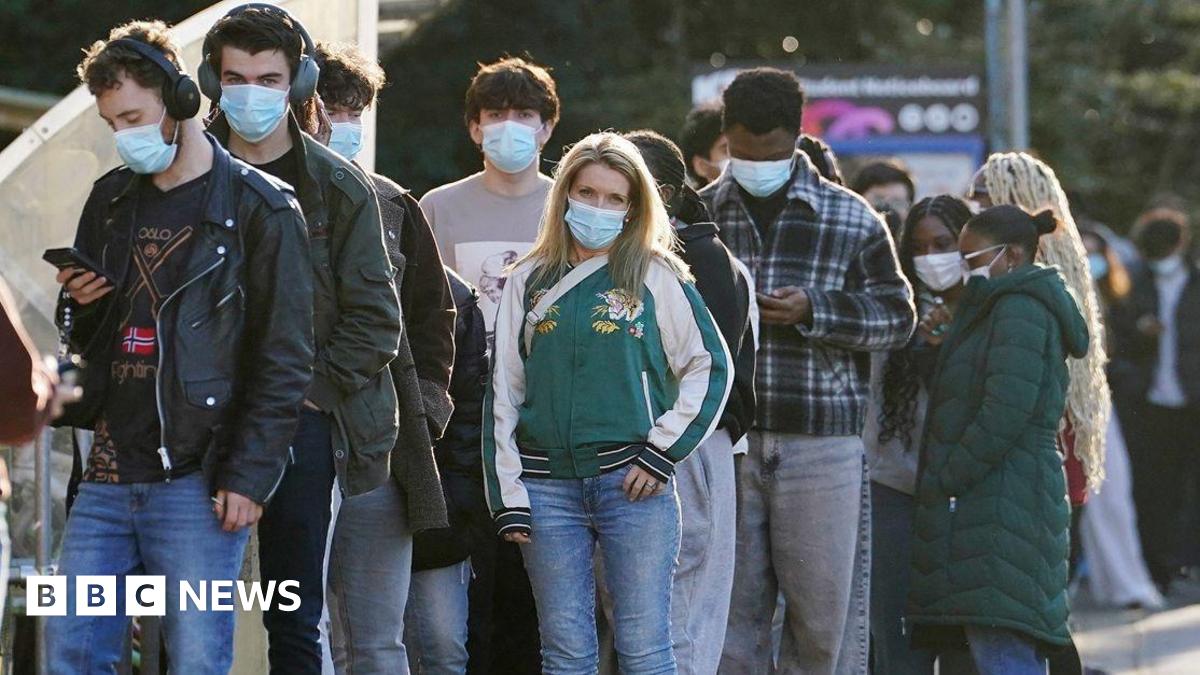 Students wait in line at the entrance to the sports hall at University of Kent campus in Canterbury. They are wearing masks and coats. Some are looking their phones, one has headphones on. Some are chatting in groups.