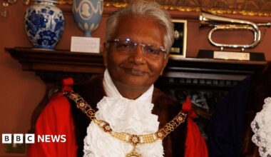Wearing his mayoral robes and gold chain, Bharat Pankhania smiles at the camera. Behind him two vases and a musical instrument can be seen on a mantelpiece.