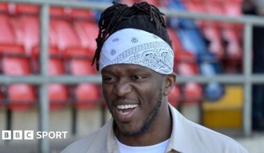 A smiling KSI wearing a beige jacket, white T-shirt and white bandana. The Dagenham & Redbridge stadium can be seen blurred behind him