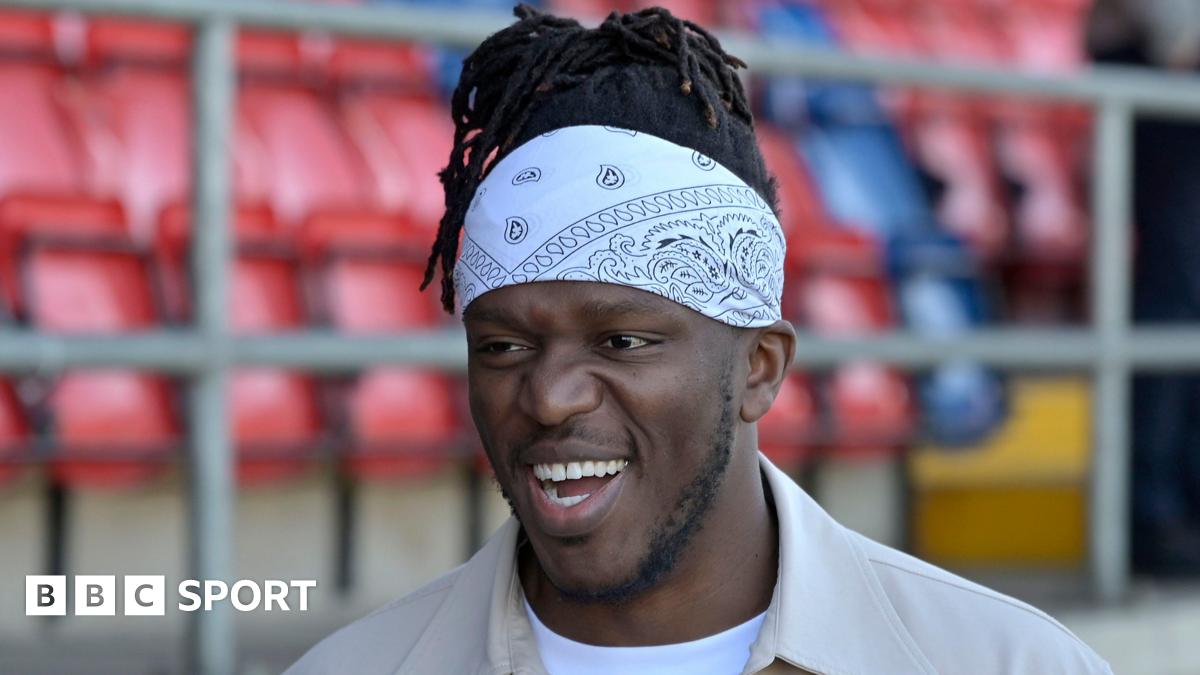 A smiling KSI wearing a beige jacket, white T-shirt and white bandana. The Dagenham & Redbridge stadium can be seen blurred behind him