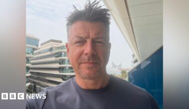 Luke Deal in a selfie-style image while he stands on a hotel balcony. He is slightly smiling at the camera. He has short dark hair with some grey. He wears a grey T-shirt.