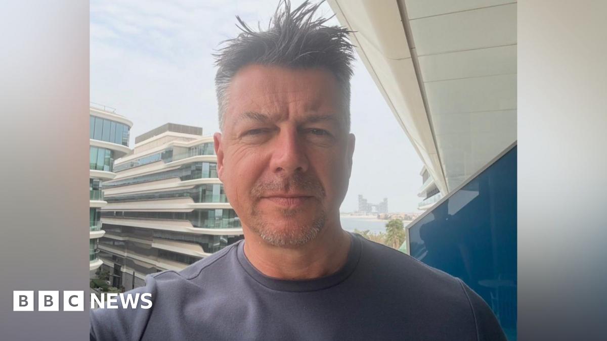 Luke Deal in a selfie-style image while he stands on a hotel balcony. He is slightly smiling at the camera. He has short dark hair with some grey. He wears a grey T-shirt.