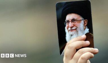 Just a hand can be seen, the rest of the person out of shot - holding up a picture of the late Iranian Supreme Leader Ayatollah Ali Khamenei. He is an elderly man with a white beard, pictured smiling. The portrait is about the size of a playing card.