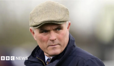 Richard Evan Rhys Williams is pictured leaving Cardiff Crown Court. He is smiling through the sunshine, squinting slightly. He has grey short hair and wears a navy suit, white shirt and navy tie.