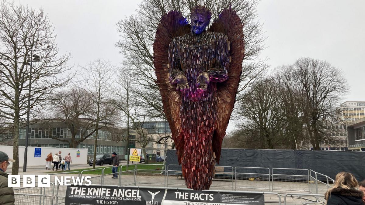 The Knife Angel sculpture in Plymouth. People are stood next to the artwork made from 100,000 blades. Metal fencing has been placed around the sculpture.