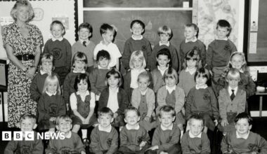 A black and white photo of Gwen Mayor, a blonde-haired woman in a patterned dress, and her 28 pupils arranged in four rows of seven in a classroom. The children are in school uniform.