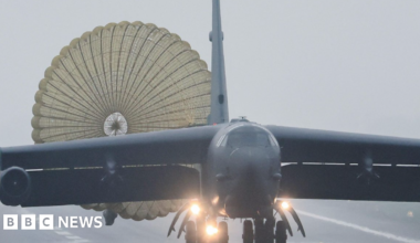 A large black B52 military bomber, a long sleek black plane, coming down the runway at Fairford air base. It is a misty day and there is barbed wire in the foreground.