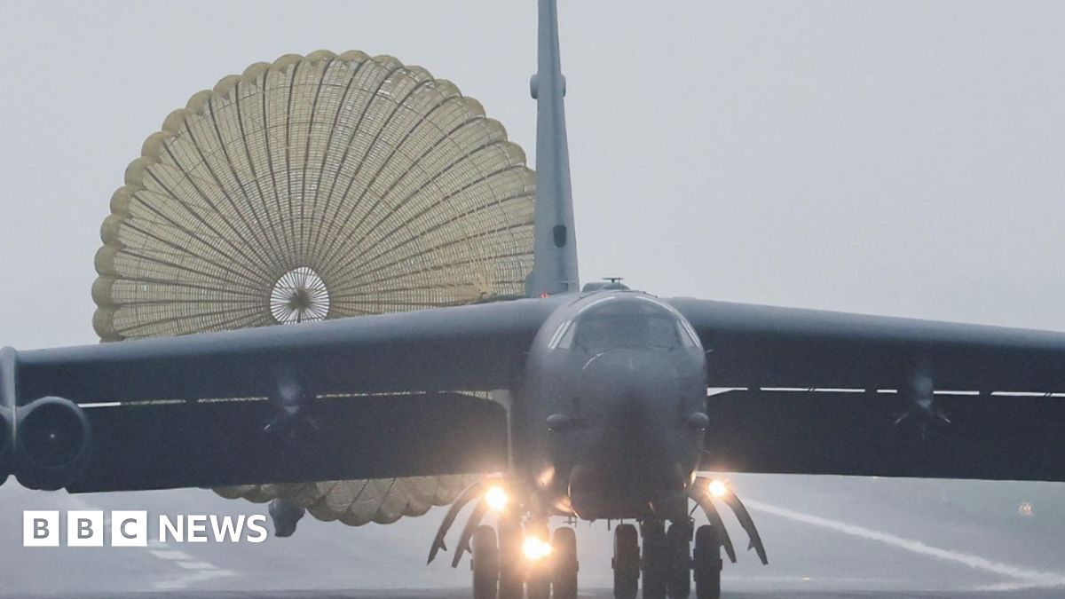 A large black B52 military bomber, a long sleek black plane, coming down the runway at Fairford air base. It is a misty day and there is barbed wire in the foreground.