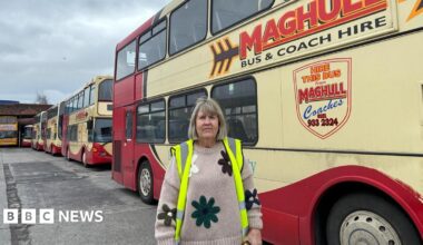 A woman with light brown hair cut into a fringe, wearing a high-vis jacket over a knitted beige jumper with a floral design, looks into the camera on a concrete bus depot with red and yellow double decker buses in the background.
