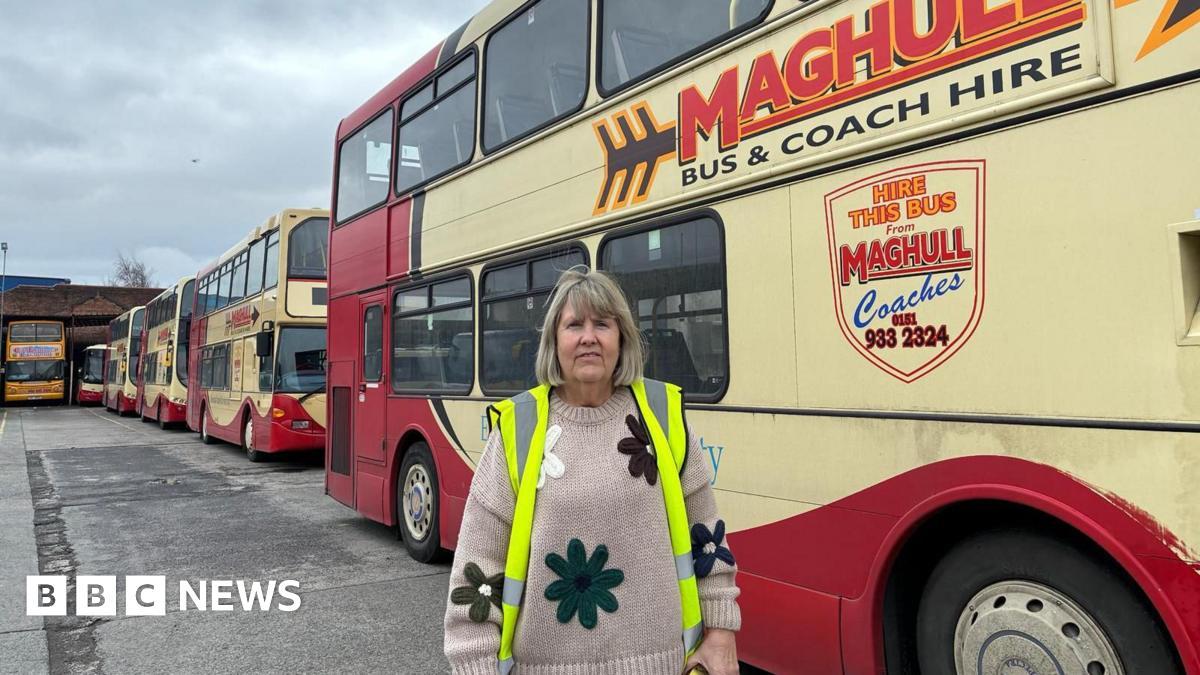 A woman with light brown hair cut into a fringe, wearing a high-vis jacket over a knitted beige jumper with a floral design, looks into the camera on a concrete bus depot with red and yellow double decker buses in the background.