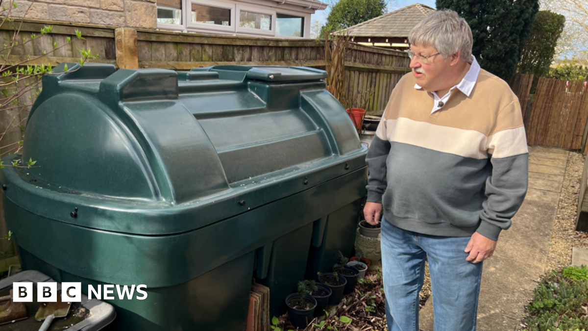 A man in a jumper stands next to a green oil tank