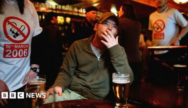 A man sitting at a table in a bar smokes a cigarette with a pint in front of him. Behind him, people are wearing t-shirts with a smoking ban symbol and the date of 26 March 2006