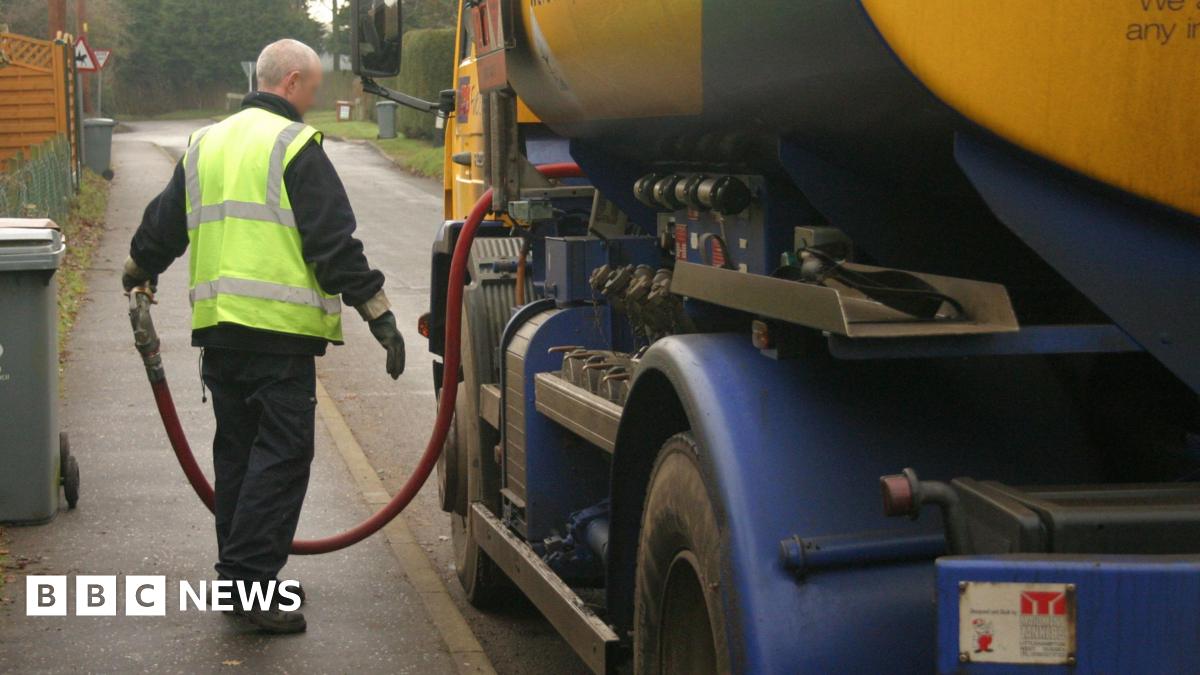 A delivery driver pulls the hose out of a small oil tanker lorry as it is parked by the side of a residential street.