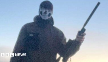 A blurry image of the boy standing in a field and holding a gun. He is wearing a mask with a skull design on it. His eyes above are blurred. He is wearing a dark waterproof jacket.