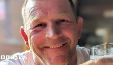 Paul Quinn, who has short light brown hair, smiles at the camera while holding a pint of what appears to be lager and wearing a white vest-top.