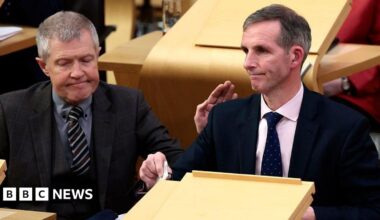 Liam McArthur, a man with greying hair, wearing a dark suit and pink shirt, sits in the Holyrood chamber looking despondent. He is being patted on the shoulder by Willie Rennie, a man with grey hair, a grey three-piece suit and grey shirt.