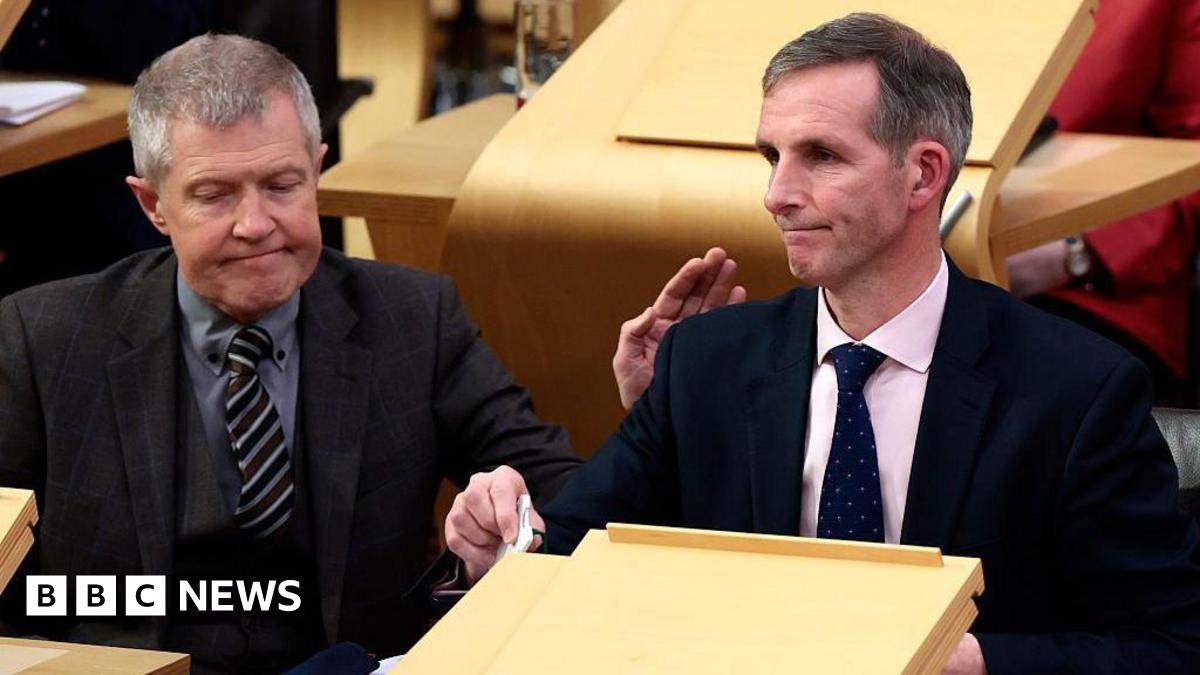 Liam McArthur, a man with greying hair, wearing a dark suit and pink shirt, sits in the Holyrood chamber looking despondent. He is being patted on the shoulder by Willie Rennie, a man with grey hair, a grey three-piece suit and grey shirt.