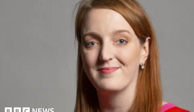 Portrait of Charlotte Nichols - Warrington North MP - with long red/light brown hair wearing a red and hot pink top. She is smiling.