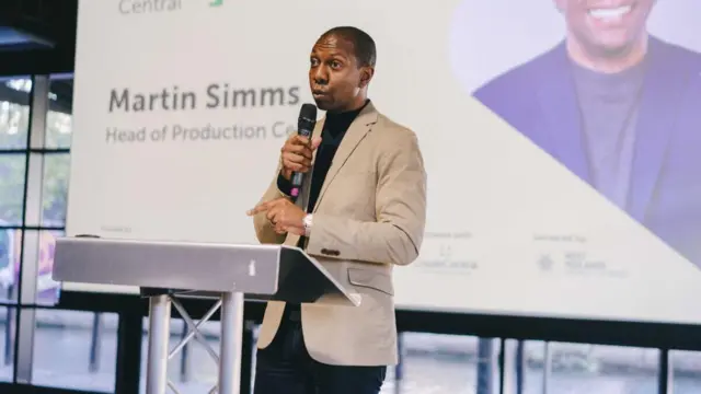 Martin Simms has short black hair and is standing on a stage at a podium with a black microphone to his mouth. He is wearing a black top with a beige suit jacket. There is a screen behind him with a photo of him on it, and it reads "Martin Simms, head of production central"