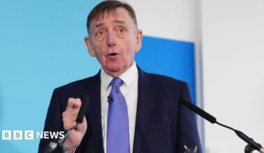Sir Robin Wales speaks into a microphone at a press conference, in front of a blue backdrop.