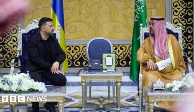 Ukraine's President Volodymyr Zelenskiy meets Deputy Governor of Makkah Region Prince Saud bin Mishaal bin Abdulaziz. The pair are seated in elaborate chairs with a silver-and-gold table between them. The room they are in is palatial, with a gold-and-black printed pattern on the walls. Zelensky is wearing an all-black outfit with a jacket, and the Deputy Governor wears a red-and-white traditional headscarf.