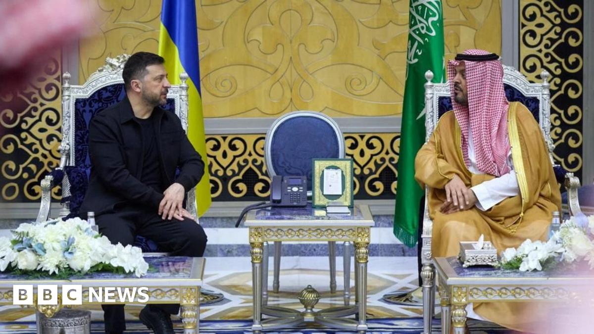 Ukraine's President Volodymyr Zelenskiy meets Deputy Governor of Makkah Region Prince Saud bin Mishaal bin Abdulaziz. The pair are seated in elaborate chairs with a silver-and-gold table between them. The room they are in is palatial, with a gold-and-black printed pattern on the walls. Zelensky is wearing an all-black outfit with a jacket, and the Deputy Governor wears a red-and-white traditional headscarf.