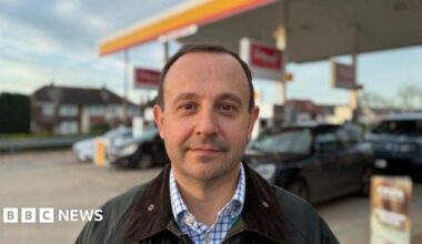 Man with short hair in open necked shirt and Barbour style jacket standing on a petrol station forecourt