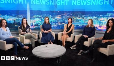 A woman, Ghislane Maxwell, with a short haircut smiles and stands behind a white podium to her left a man, Kevin Spacey, has a cell phone on his right ear. He is wearing a navy shirt and blue jeans.