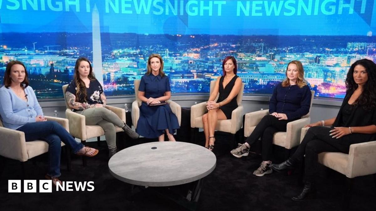 A woman, Ghislane Maxwell, with a short haircut smiles and stands behind a white podium to her left a man, Kevin Spacey, has a cell phone on his right ear. He is wearing a navy shirt and blue jeans.