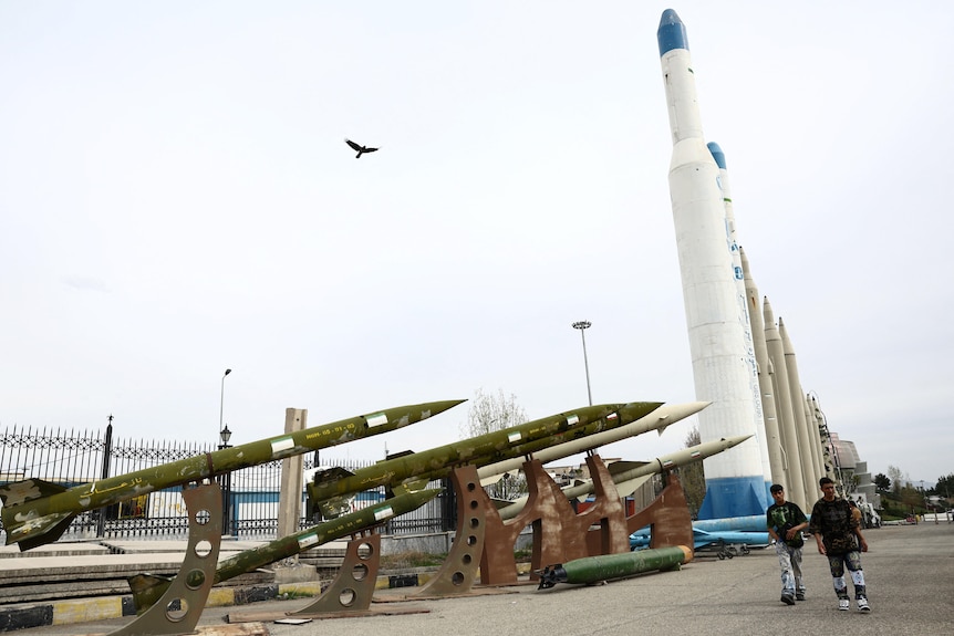Two people walking on the street with large missiles lined up on the street
