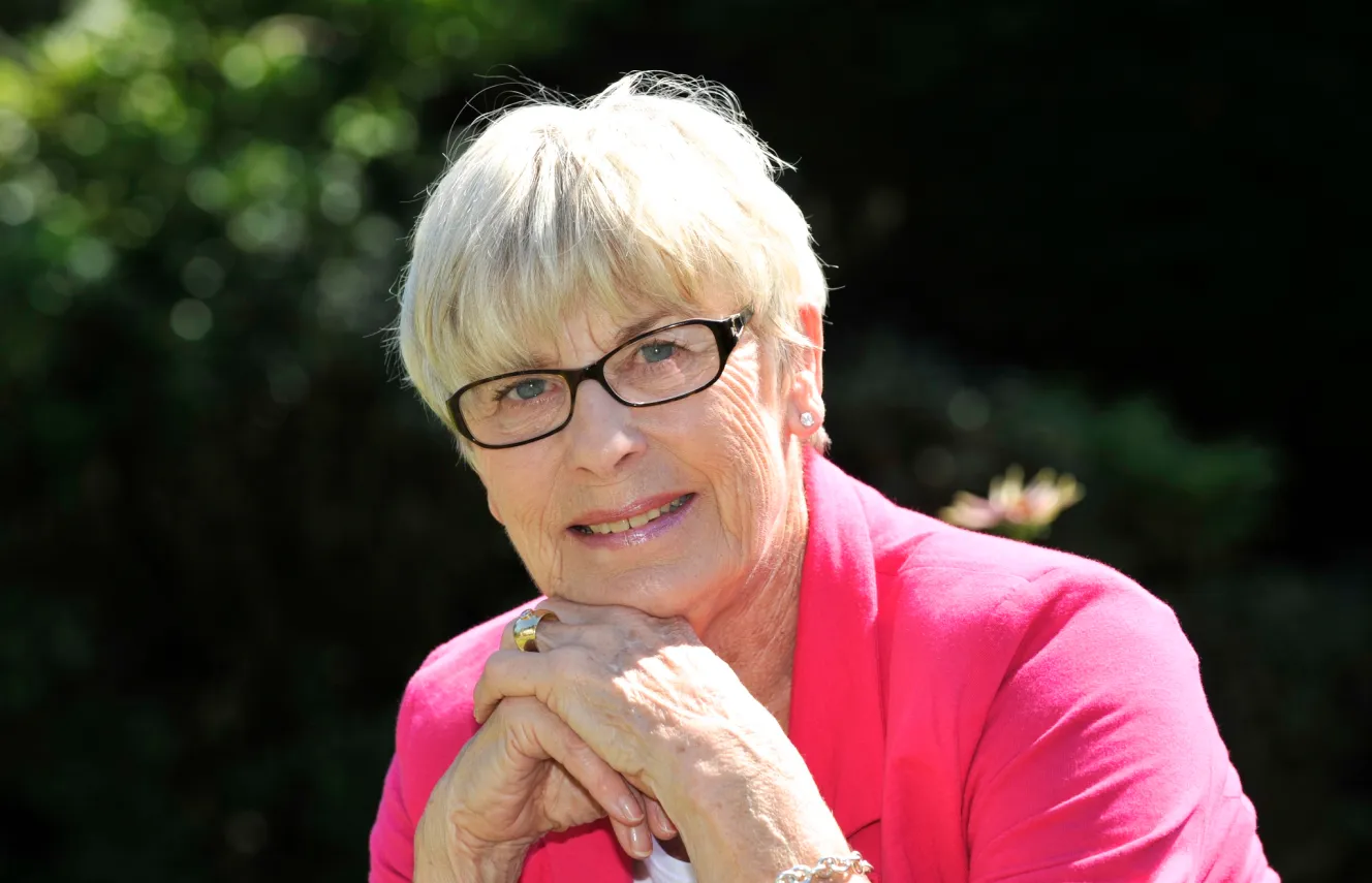 Ex Olympian Mary Rand wearing glasses and a pink top, smiling with her hands clasped under her chin.