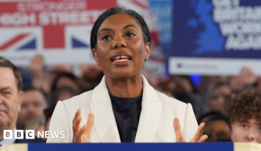 Kemi Badenoch wearing a white jacket and a black top standing in front of a crowd of people some of whom are holding up placards