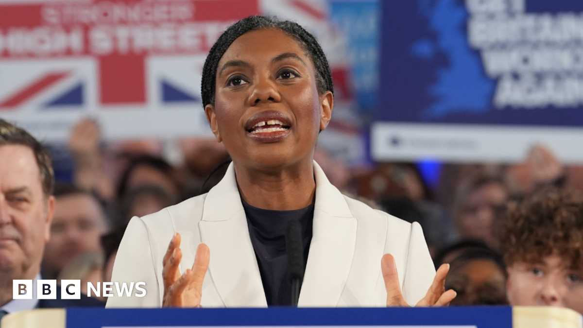 Kemi Badenoch wearing a white jacket and a black top standing in front of a crowd of people some of whom are holding up placards