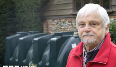 David Coverdale is wearing a red zip-up jumper over a checkered shirt. He has grey hair and a goatee. Behind him is a large dark green oil tank.