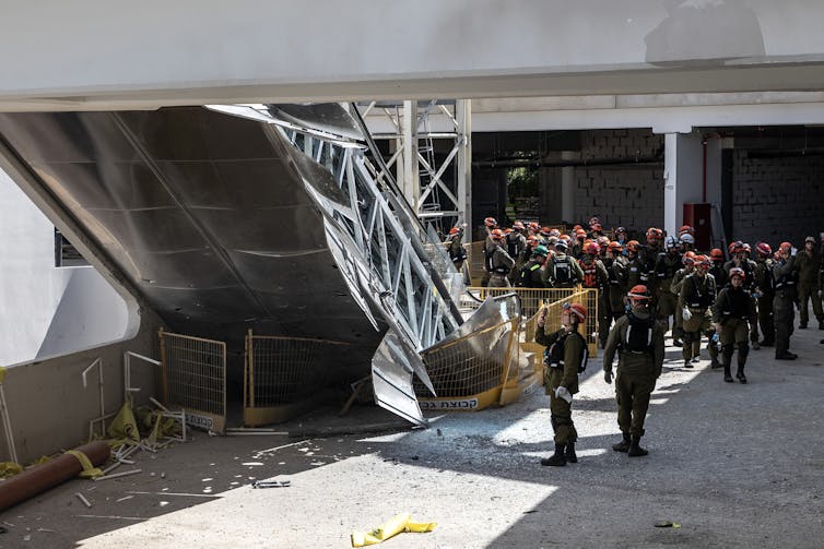 A group of people wearing uniforms gather near a large steel, collapsed structure that appears to be part of a bridge