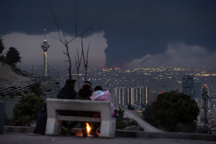 Three people sit on a bench and look over a city that has dark smoke rising from it.