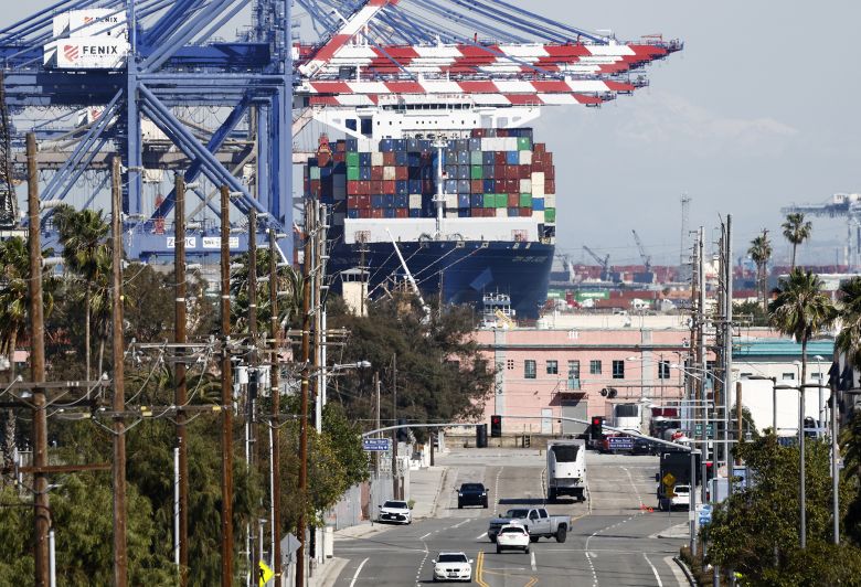 A container ship floats loaded with shipping containers at the Port of Los Angeles as vehicles drive nearby on February 20, 2026.