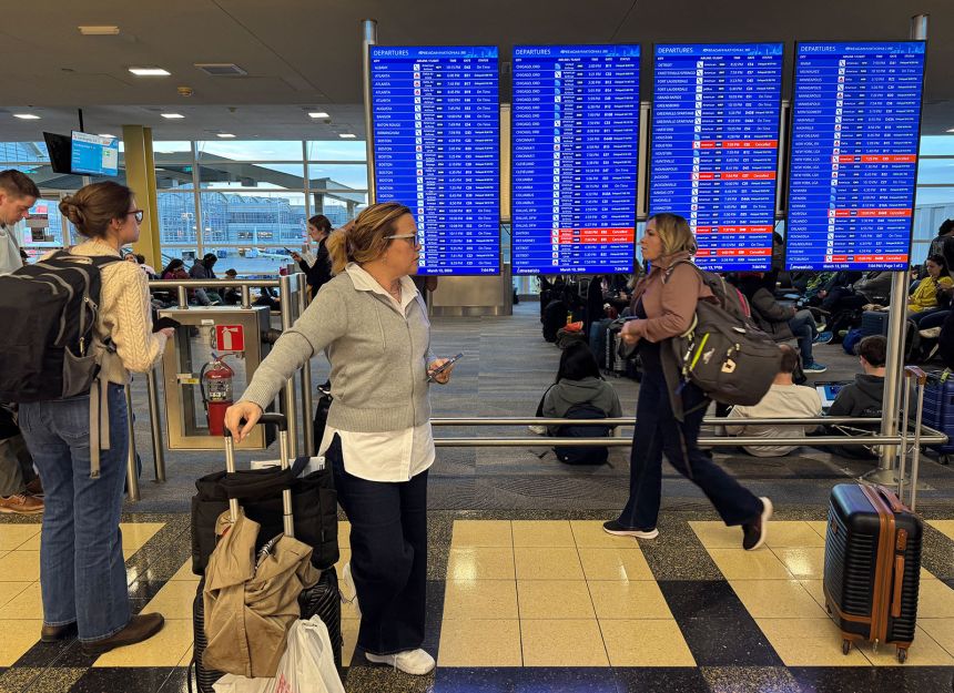 Travelers walk past a departures screen inside a terminal during a ground stop at Ronald Reagan Washington National Airport in Arlington, Virginia, on March 13, 2026.